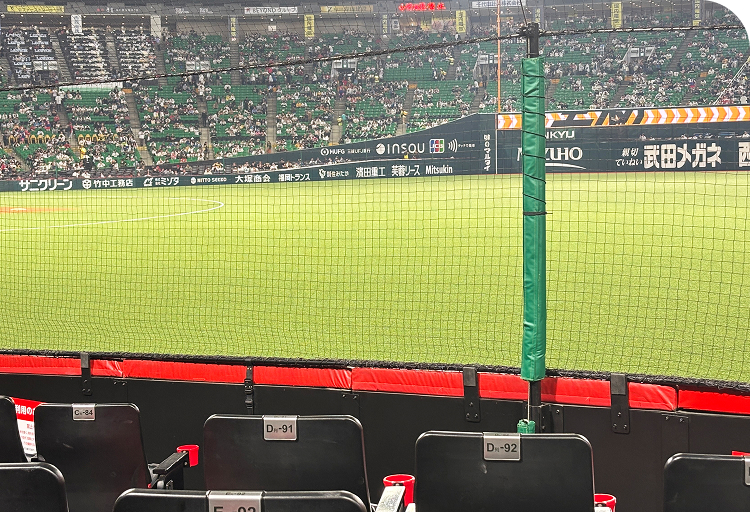 観客席からネット越しに、緑の芝生が広がる野球場のフィールドを眺めています。